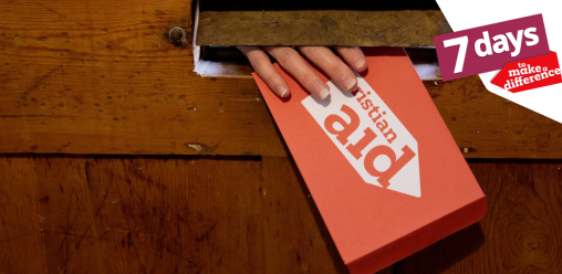 A hand reaching through a letter box, holding a red Christian Aid envelope.