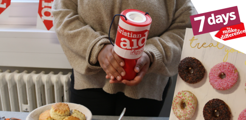 A woman holding a Christian Aid donation bucket, stood behind a table full of baked goods. There is Christian Aid bunting behind her.