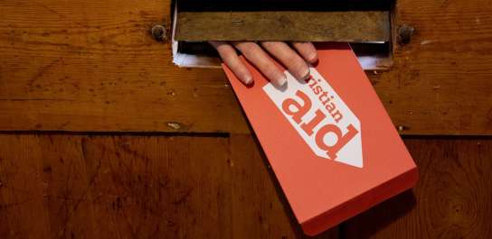 A hand reaching through the letterbox, holding a red Christian Aid envelope.