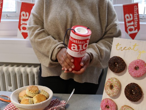 A woman holding a Christian Aid donation bucket, stood behind a table full of baked goods. There is Christian Aid bunting behind her.