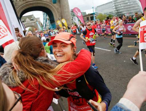 Two women hugging. One is wearing a cap and is running along a marathon track, the other is supporting her from the crowd. There are Christian Aid flags around, and many people are wearing red Christian Aid shirts.