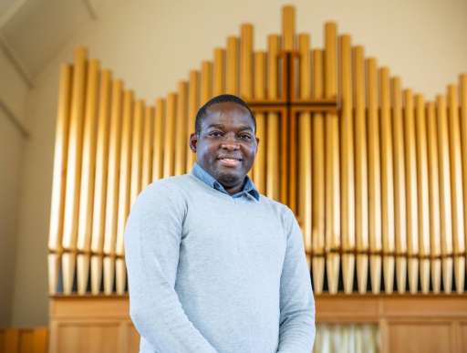 A man stands in a church smiling at the camera. Behind him is the pipes of an organ
