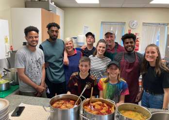 A group of young people smiling at a Christian Aid Week fundraising event. They stand behind a counter with various pots of food.