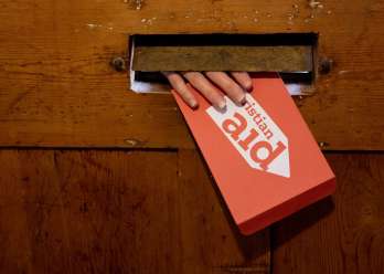 A hand reaching through the letterbox, holding a red Christian Aid envelope.