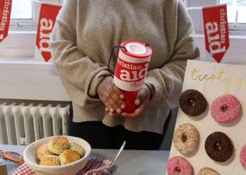 A woman holding a Christian Aid donation bucket, stood behind a table full of baked goods. There is Christian Aid bunting behind her.